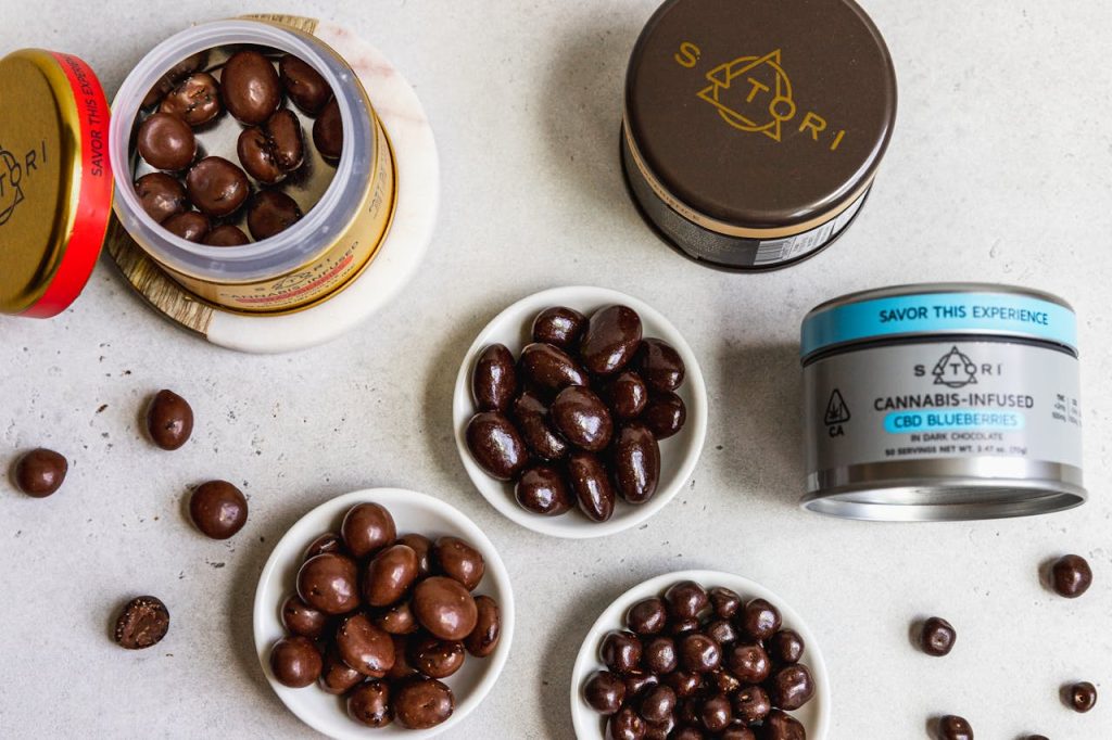 Satori cannabis-infused CBD blueberries in dark chocolate, shown in tins and small white dishes on a gray surface.